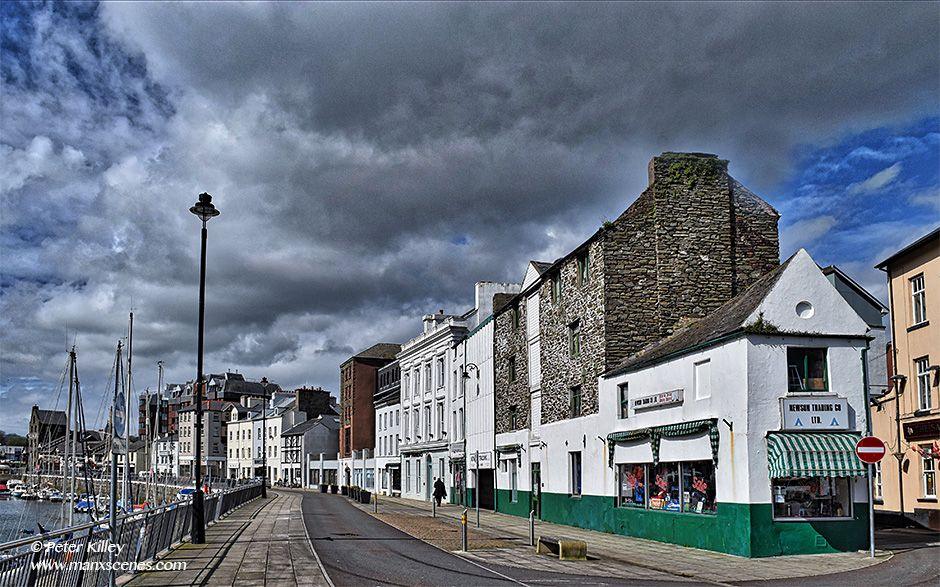 An old photo of Douglas Quayside, the site has a small white building with green paint on the bottom, which goes into a taller stone building, with white and green paint on the bottom.