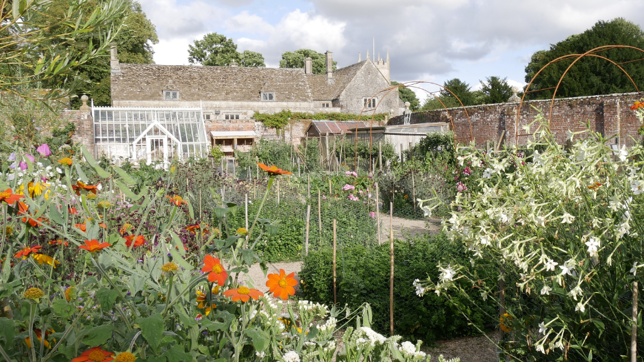 The kitchen garden in the summer with flowers and vegetables blooming in the sunshine, with a greenhouse and the manor in the background
