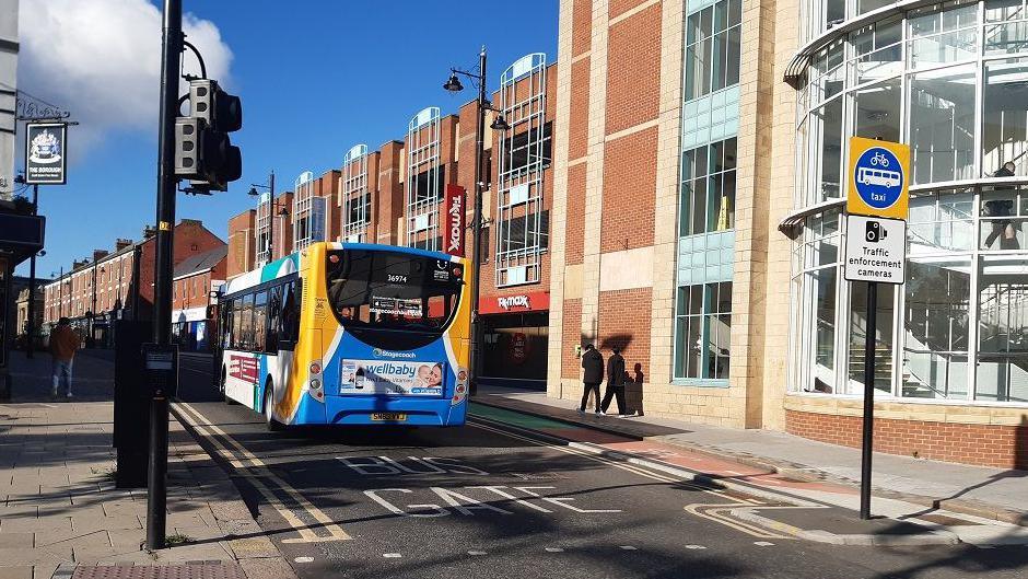 A single-decker bus driving down a street. 'Bus Gate' is written on the road and a sign with 'traffic enforcement camera' alongside a picture of a bike, bus and the word 'taxi'. Black cameras are pointing down the street.