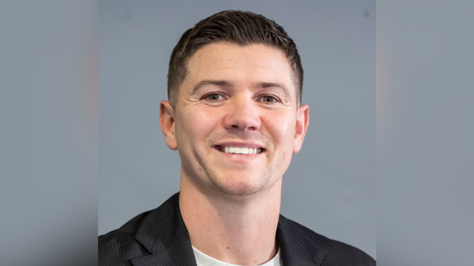 A man with brown hair smiling. He is wearing a black jacket with a white top underneath it. He is in front of a grey background.