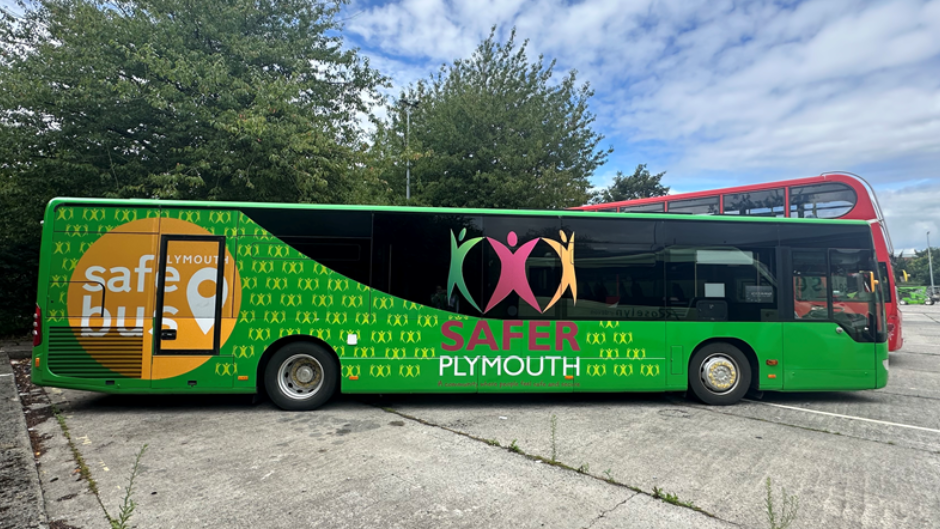 A view of the side of the bus. The bus is green and has stickers promoting Safer Plumouth