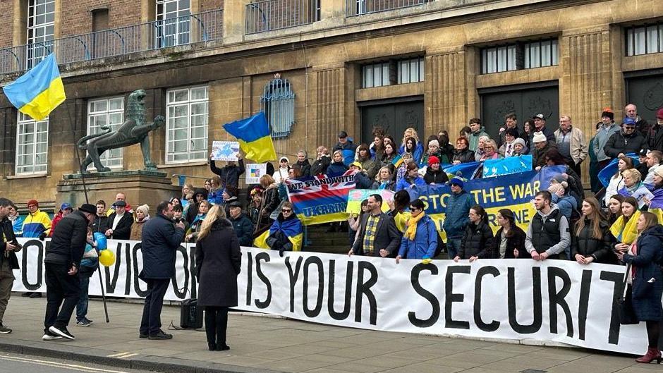 People are standing on the steps which lead up to City Hall in Norwich. Many are holding Ukrainian flags. A speaker holding a microphone is standing in front of them.