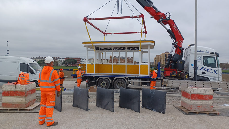 A yellow, white and blue shelter being lowered from the back of a lorry with workers in orange hi-vis watching the operation