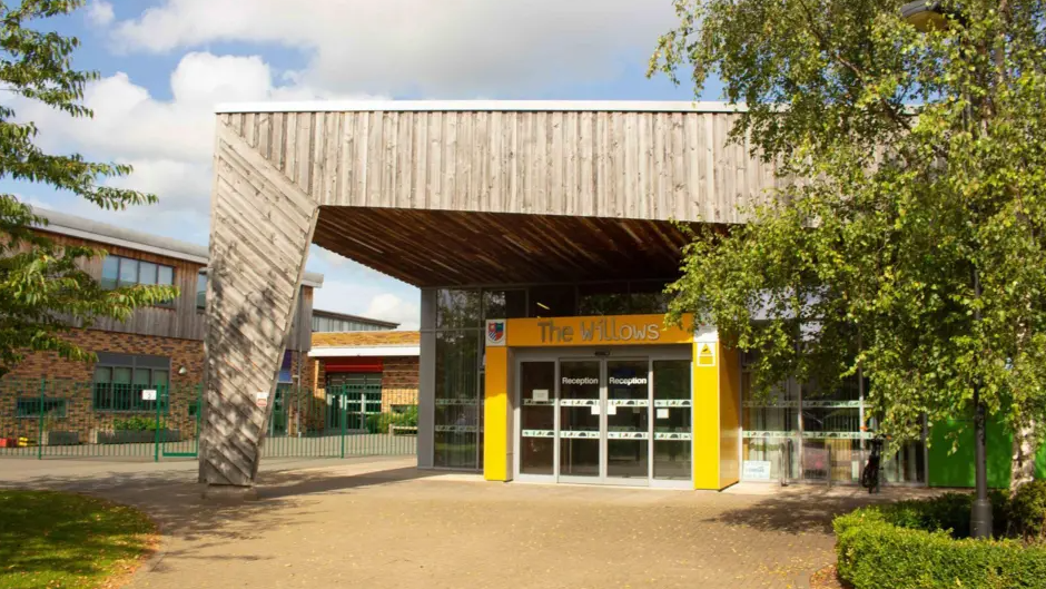 The school has a large wooden facade which spans the entrance and provides cover with a long piece of wood in keeping with the overall design touching the ground away from the entrance. The entrance is decorated yellow with glass doors and windows.