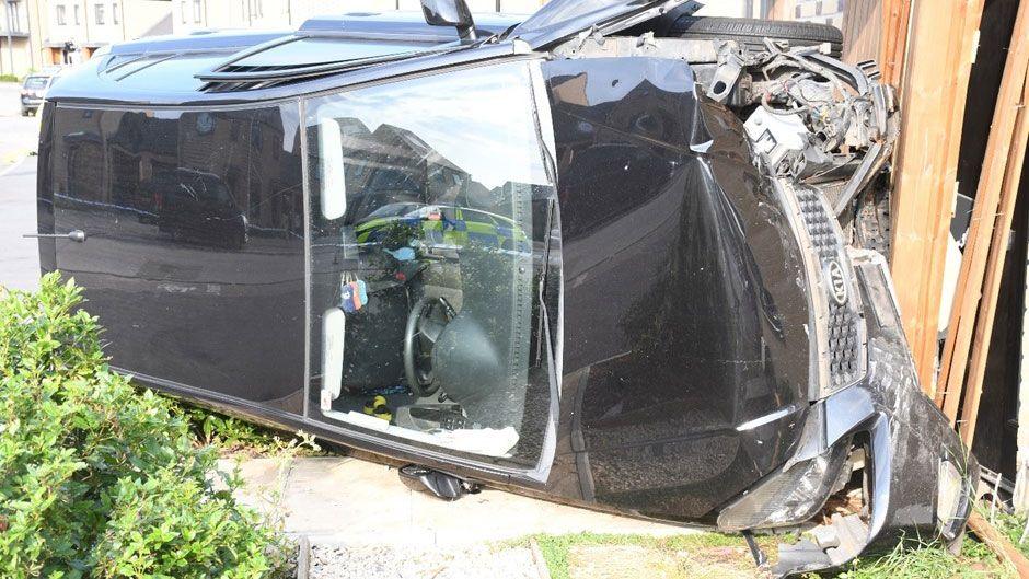 A black Kia car resting on the driver's side after it appears to have crashed into a house. The front of the car is all smashed up and the passenger door is slightly open.