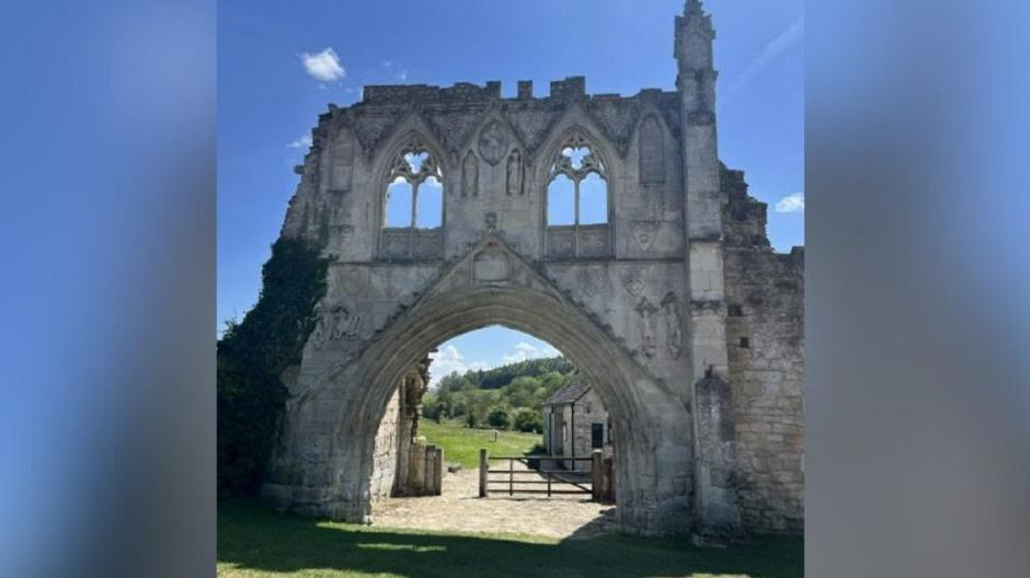 The remains of an Augustinian priory at Kirkham Abbey which is made up of cream coloured stones and has an archway leading to fields and countryside.