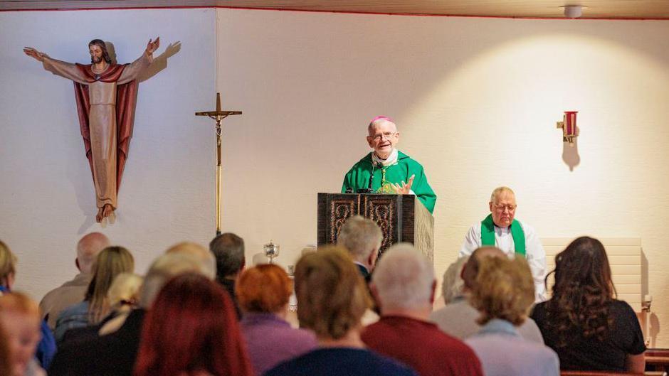 A bishop stands at the front of a church, wearing a green cassock and a purple skull cap. A priest in a white robe and green stole (scarf) sits beside him. A cross is to the left of the picture and there is a mounted statue of Christ with open arms on the far left. In the foreground are the backs of heads of people who are attending the service.