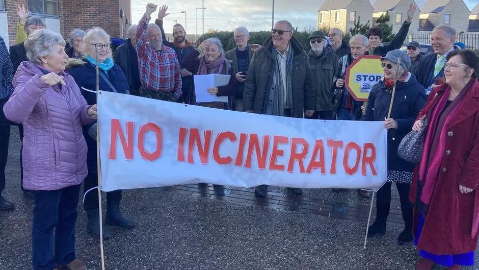 A group of about 15 people standing with a banner saying "no incinerator" in red, capital letters on a white background