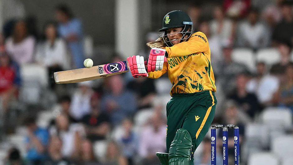 Suné Luus looks to crack the white cricket ball through the on-side, wearing green and gold South Africa kit and red and white batting gloves, with shoulder length blonde hair pretruding from bottom of her helmet
