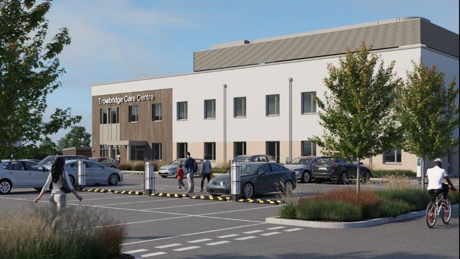 A computer generated image of what the care centre will look like once finished. It is a large white building clad with the entrance block clad in wood. There is a car park in front with people walking and cycling across it.