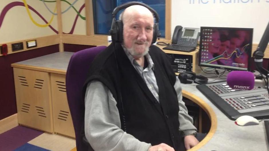 John Kaneen sitting in a Manx Radio branded radio studio. He is wearing headphones, a grey shirt and black wool waistcoat. There is a microphone, telephone, radio studio desk with lots of buttons and a computer screen near by.