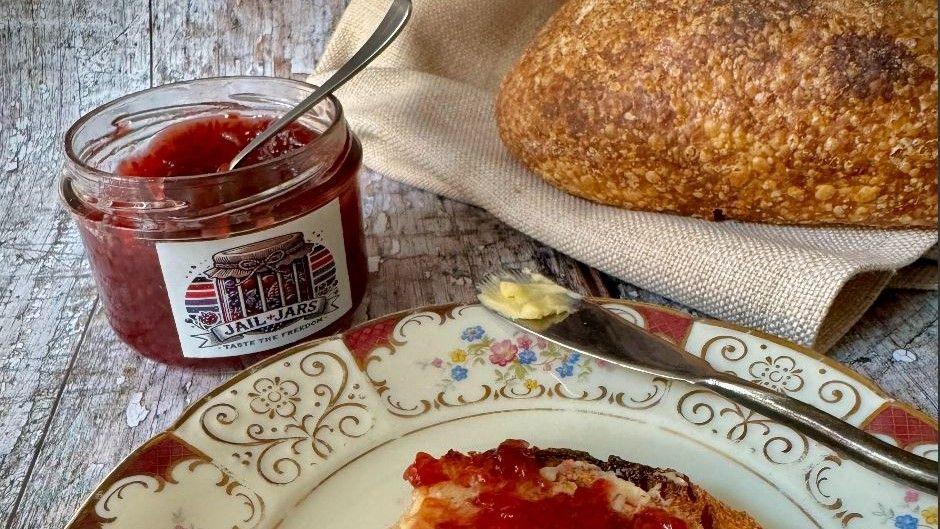 A jar of jam on a wooden table with a label on it which says Jail Jars. Beside it there is an ornate china plate with a piece of jam toast on it and a butter knife resting on the side. Also on the table is a crusty loaf of bread laying on a burlap sack. 