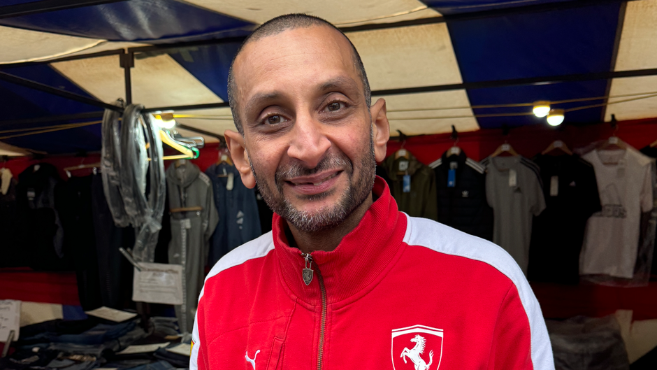 Bob Sidhu smiling. He is wearing a red jacket. He is in a market and their are clothes behind him.