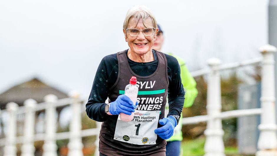 An elderly woman with short blonde hair and glasses runs a previous Hastings Half Marathon in the rain with a smile on her face in a black and green Hastings Runners uniform