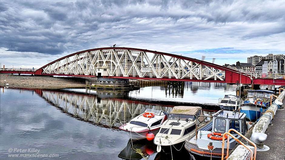 The bridge an arching red structure with cream metal rails connecting them, boats are in a harbour in the foreground.