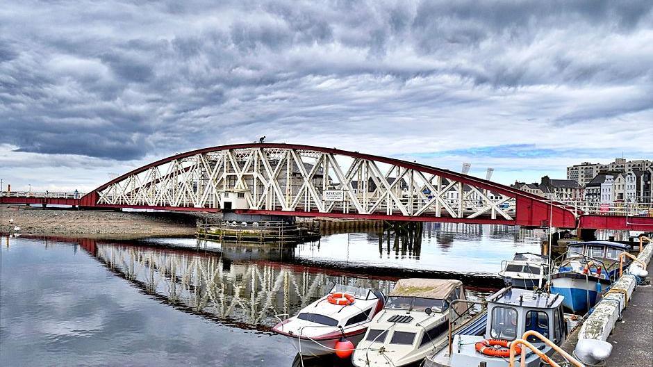 The Ramsey Swing Bridge, which is painted red and white. It's reflection can be seen in the water below it alongside small pleasure boats moored against the harbour wall. The sky above is filled with dramatic grey clouds, with a hint of blue sky in the distance.