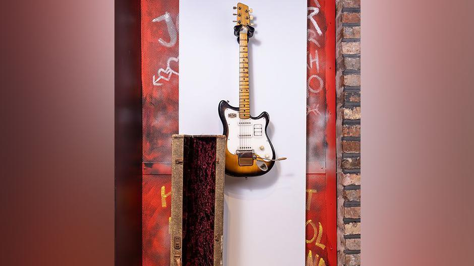 George Harrison's Futurama electric guitar hanging up on a white board on a wall along with an old guitar case and the red wall littered with graffiti 