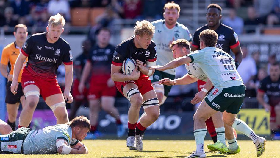 Nathan Michelow (centre) bends his knees and holds the ball close to his body under an arm as two Leicester players come towards him