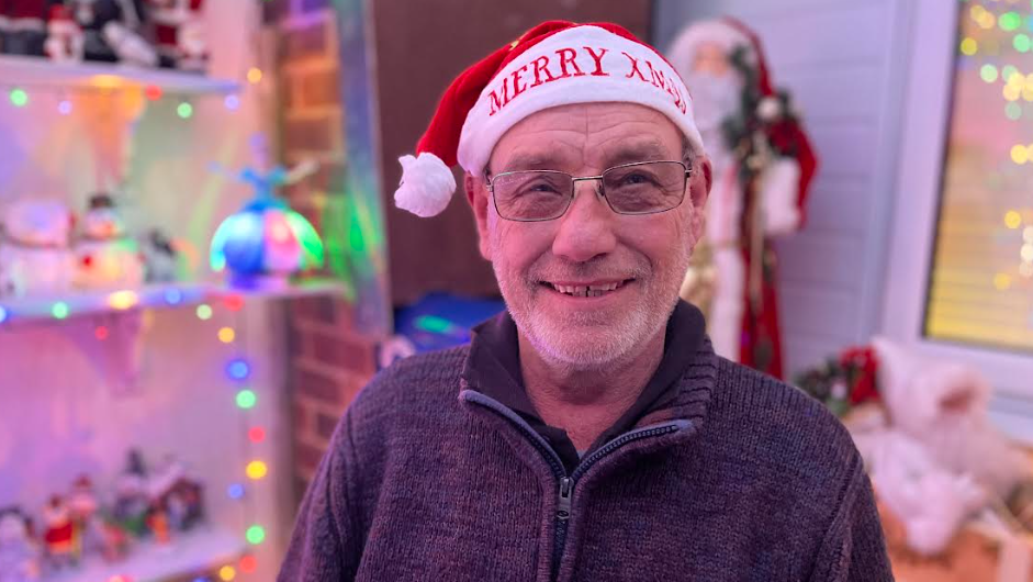 A man wearing glasses and a Santa hat smiles at the camera. Multicoloured fairy lights are visible in the background 