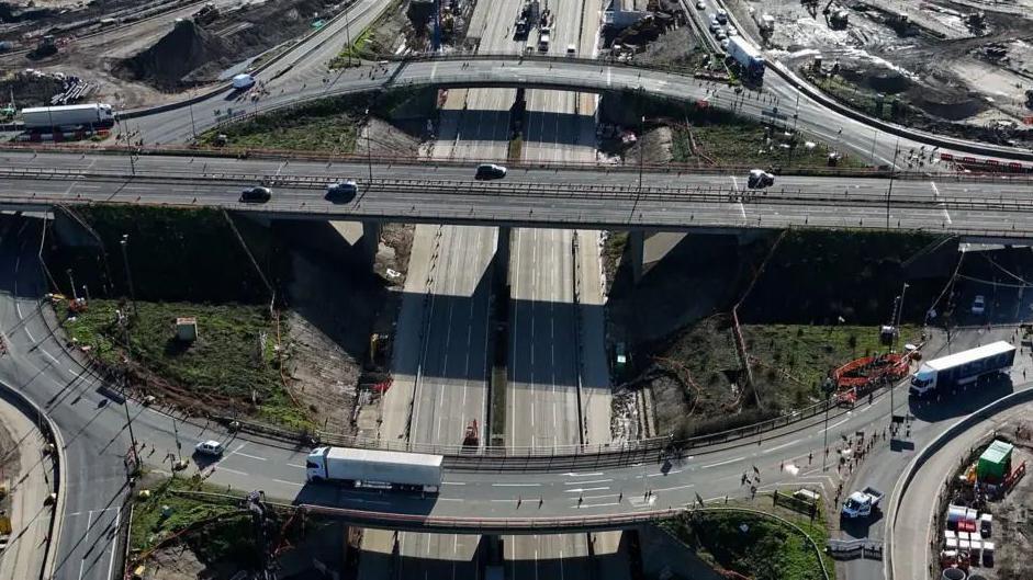 Junction 10 of the M25 seen from above. There are a number of vehicles driving on the roads, and an area of grass in the middle.