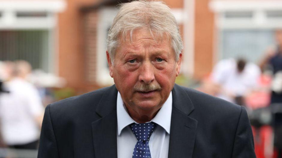 A man looks into the camera. He has white hair and a white and brown moustache . He is wearing a dark suit, white shirt and navy tie with white spots. he background is faded and people can be seen standing in front of a red bricked property.