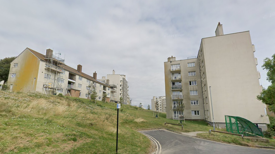Two blocks of flats. The flats are cream covered with brown dirt marks on the exterior. They are at the end of the road at the top of a small grassy hill.