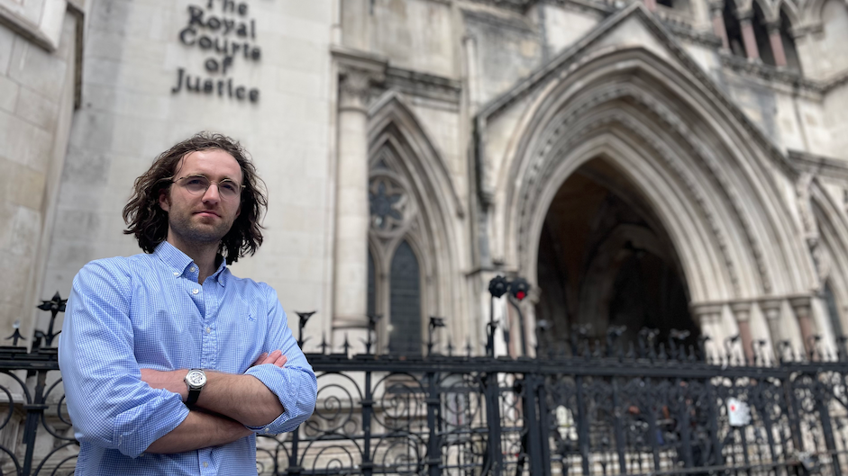 David Hamon has shoulder-length brown hair, is wearing glasses and a light blue shirt with the collar unbuttoned and has his arms crossed as he stands in front of the Royal Courts of Justice in London