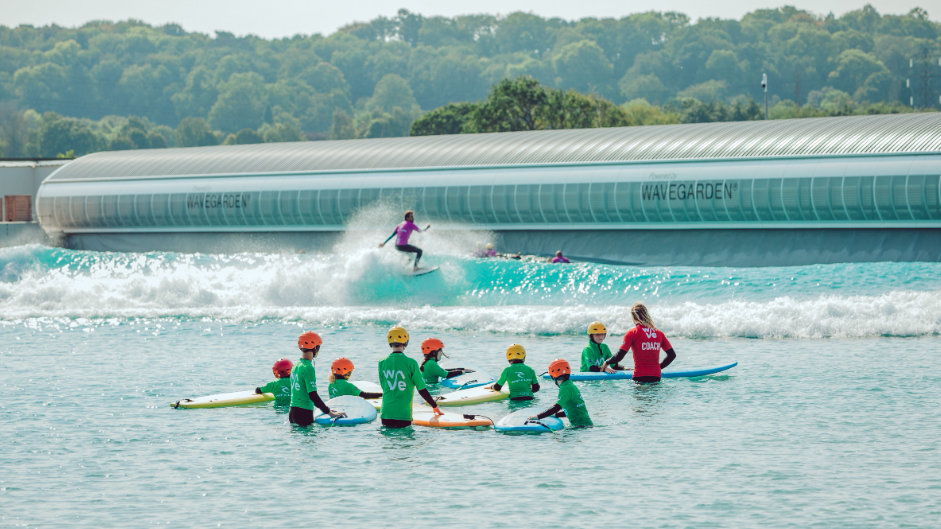 A group of children wearing helmets and green t-shirts stand in the water with a surfboard each, surrounding an instructor wearing a red t-shirt. 