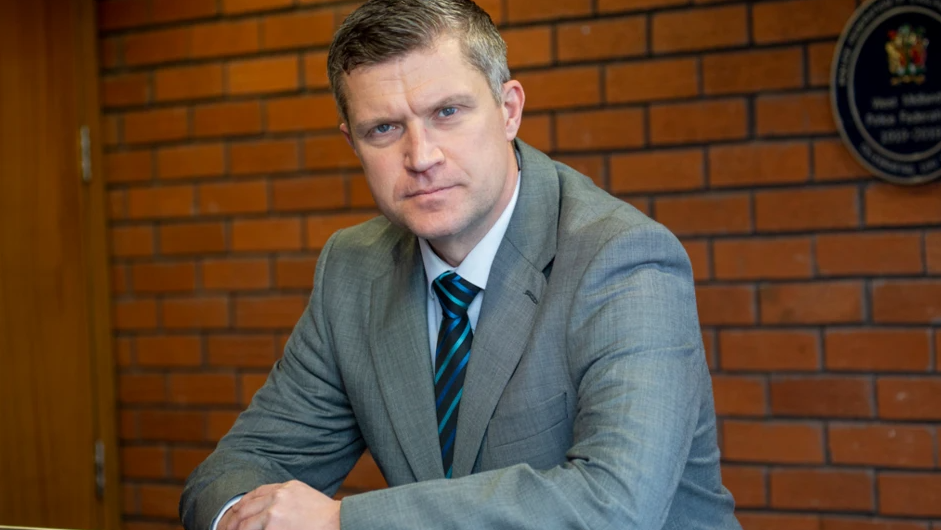 Mr Cooke is wearing a grey suit and tie and leaning on a railing inside a building with a brick wall behind him.