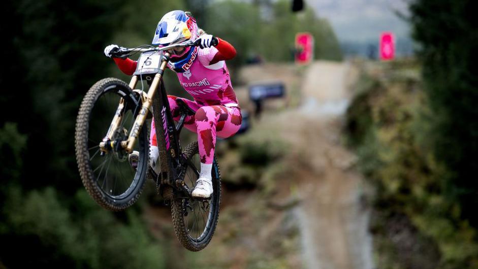 A mountain biker wearing pink clothing takes to the air from a jump at Nevis Range