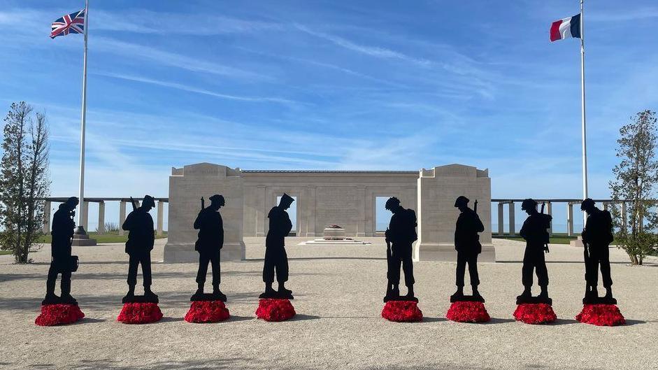 Eight figures standing in front of the D-Day memorial in Normandy. There are poppy wreathes in front of each one