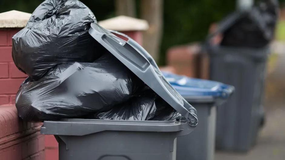 Grey bin overflowing with filled black bin bags.