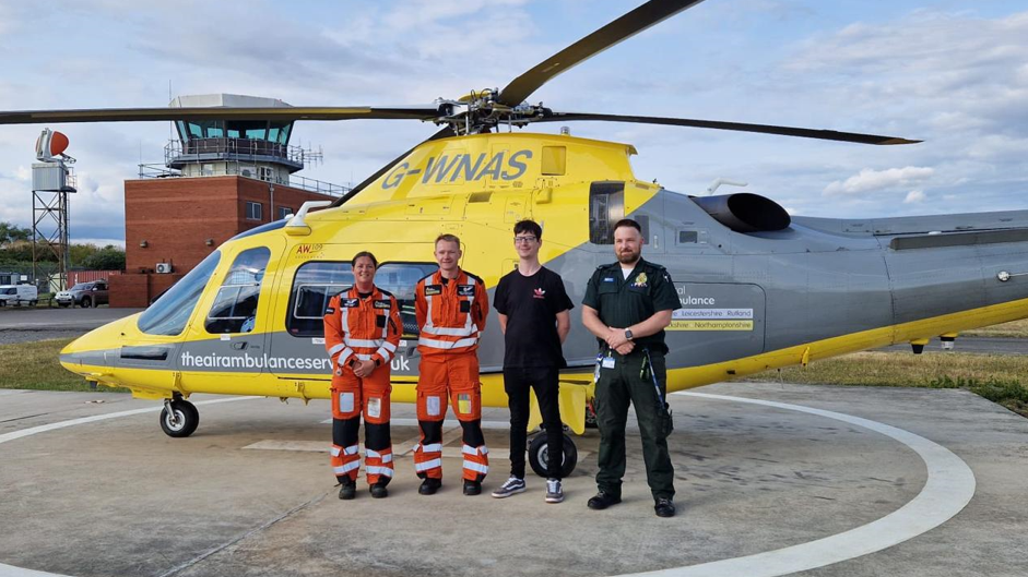 Four people, two in orange flying suits, one in a green ambulance uniform, stand in front of a yellow and grey helicopter at an airbase