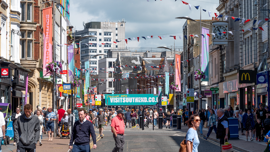 A High Street showing a buzz of activity, people walking, the sun is shining, shops are colourful and busy.