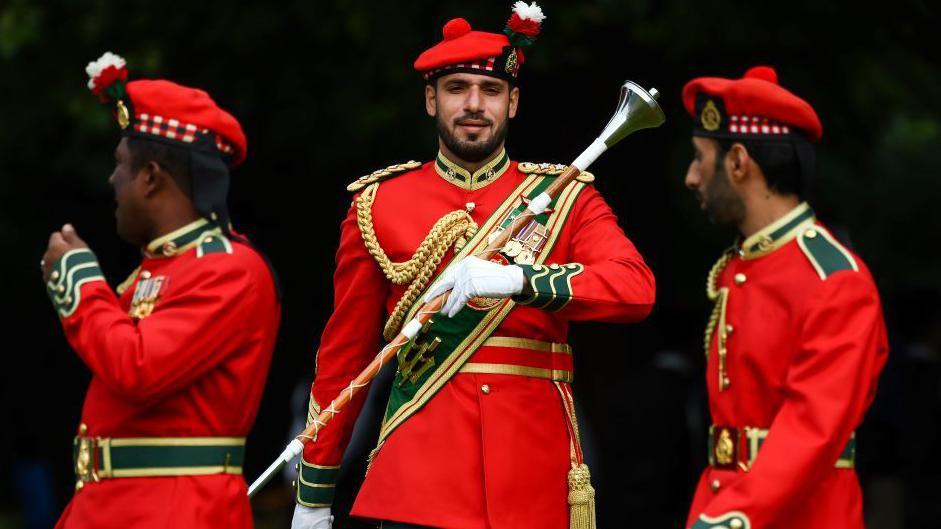 Three members The Royal Army of Oman Pipes and Drums practise., They are wearing red jackets and glengarry's. The man in the centre is holding a drum majors baton.