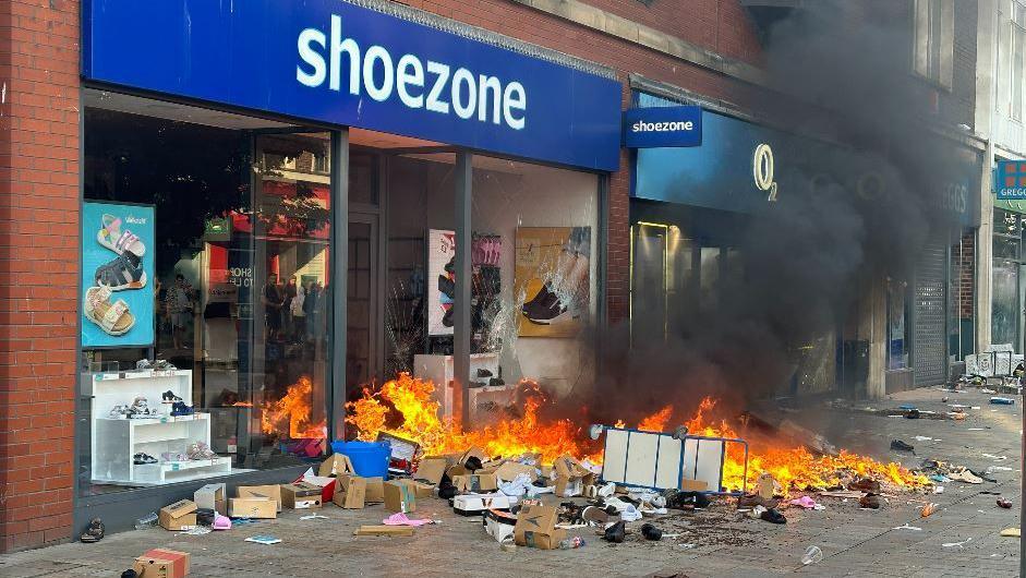 Shoezone shop with flames coming out of the doorway and boxes of shoes on the pavement. Black smoke billows into the air.