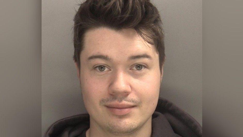 A mugshot showing the face of a young man with short dark hair and a dark hoodie against a grey background.