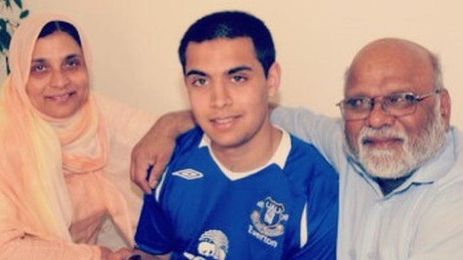 A smiling woman wearing a white top and peach coloured headscarf and a bearded man with glasses wearing a blue shirt hug a young black-heared boy wearing with a blue Everton football top 