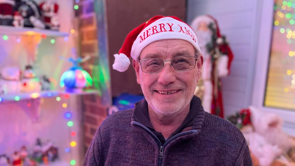 A man wearing glasses and a Santa hat smiles at the camera with multicoloured fairy lights behind him