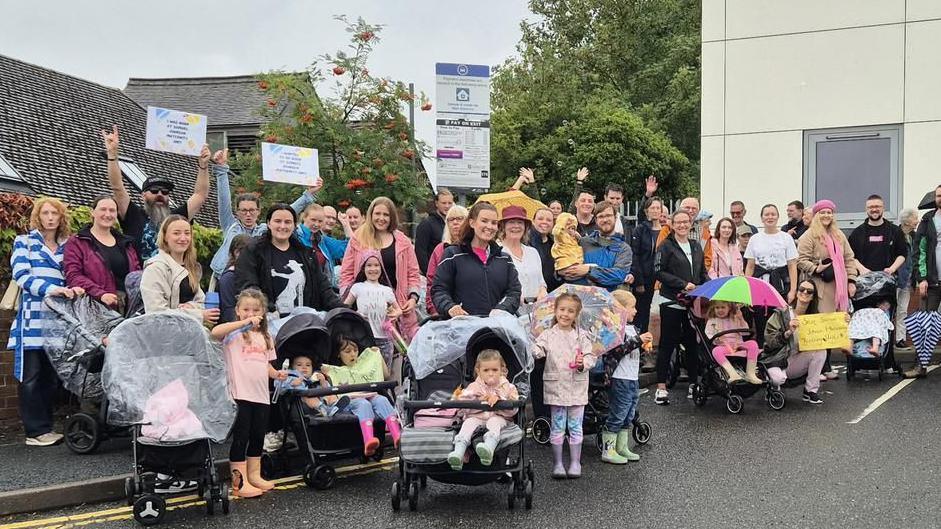 A large group of protesters including pregnant mums, families all pushing prams in the rain and smiling at the camera