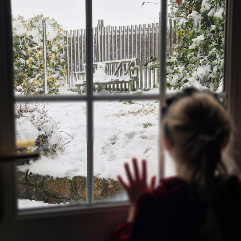 Young girl looking through a window with one hand on a pane, with snow and a small snowman on a seat outside.