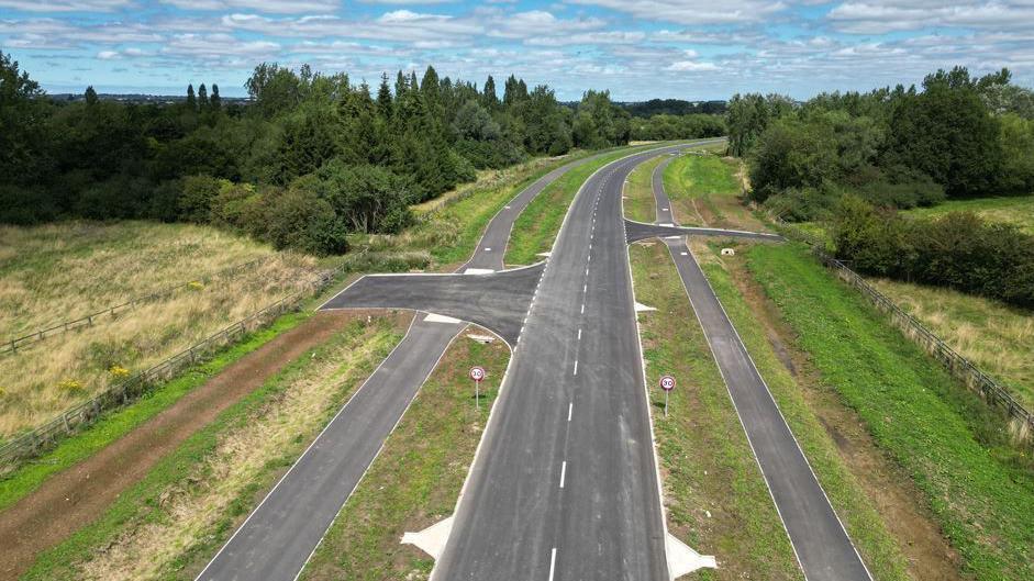 An empty grey two-lane Tarmac road in a countryside setting, with trees at the edges and grass by the roadsides. There is a footpath running either side of the road and speed signs limiting drivers to 30mph.