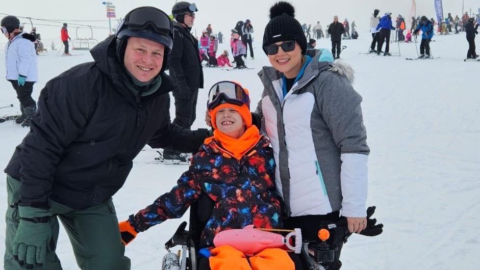 Brooke Hacker and her parents on a ski slope. Brooke is in a wheelchair wearing a blue, black and orange coat and bright orange hat, scarf and trousers. She has black goggles on her head. Her mother, Rebecca is on her right in a grey and white jacket and a black hat on her head and sunglasses. Brooke's father, Andrew is on her left, in a black coat, black helmet and green trousers.