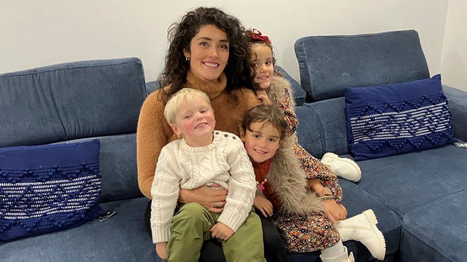 Holly Morgan is sitting on a blue coach surrounded by her three young children. They are all smiling brightly. Mrs Morgan has long black, curly hair and is wearing a brown jumper.