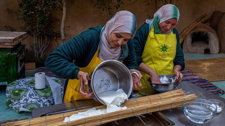 Two Egyptian women pour a cheese mixture over a wooden mat to prepare it at a community centre in Giza, Egypt - Wednesday 12 February 2025