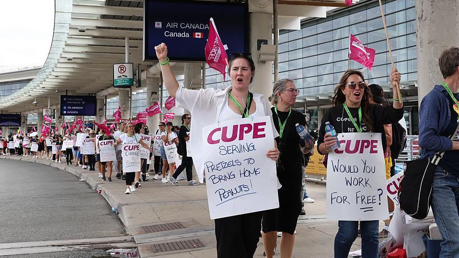 A photo showing Air Canada flight attendants picketing outside Toronto Pearson International Airport. The photo shows a long line of picketers holding signs, with a focus on two at the front with signs that read "serving pretzels to bring home peanuts" and "would you work for free?". Others are seen waving pink CUPE flags - the union representing Air Canada cabin crew.