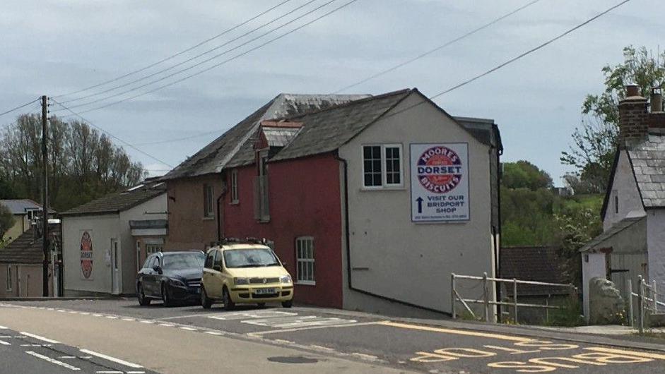 A street view of the former Dorset knob factory. It is a row of four properties in varying heights, painted red, cream and brown on the front and with a grey tile roof. On the side is a sign for the factory which has a circular logo for the biscuits on it, it reads 'Moores Dorset Biscuits. Visit out Bridport shop'. There are cars parked out the front of the building and a section of the road can be seen to the left of the image.