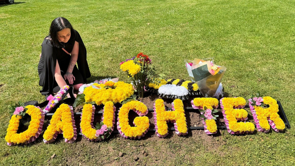 Mrs Black at a graveside with a large yellow floral wreath spelling out the word 'daughter'