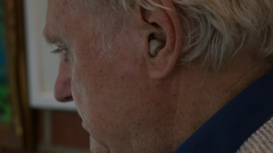 A side view of Eddie Yarrow where you can see his small white hearing aid. He is looking down and appears pensive.