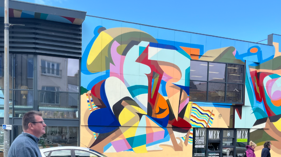 A mural featuring an abstract and vibrant composition with bold, overlapping geometric shapes and colourful patterns. The modern design is made up of angular and curved formations in a variety of bright colours, including reds, blues, yellows, pinks, and greens